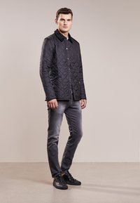 Casaco acolchoado preto com colarinho, fecho à frente por botões e bolsos laterais. Combinado com jeans cinzentos e sapatilhas pretas. Textura suave.