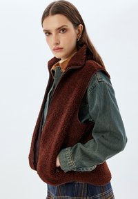 Braune Fleece-Weste mit hohem Kragen, Reißverschluss und seitlichen Taschen, über einem Jeanshemd getragen. Strukturierte Oberfläche mit dunkler Borte.