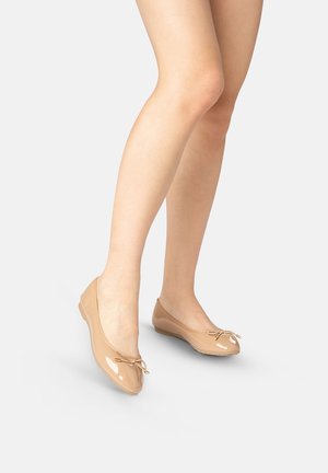 Piernas desnudas de mujer con bailarinas de charol beige con pequeños lazos, de pie sobre un suelo gris claro.