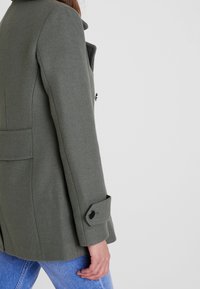 Abrigo de mezcla de lana verde con una silueta estructurada, que presenta un cuello con muescas, bolsillos laterales y detalles de puños abotonados. Llevado sobre jeans azules.