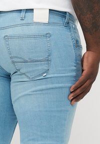 Jeans de color azul claro con un diseño ajustado, que cuentan con bolsillos traseros con costuras curvas y una etiqueta de marca en la cintura. Textura de denim suave.