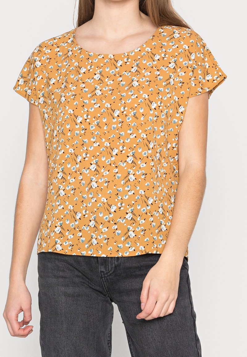 Blomstret bluse i sennepsgul med små hvide og blå blomster. Korte ærmer, rund halsudskæring og let materiale. Parret med mørke jeans.