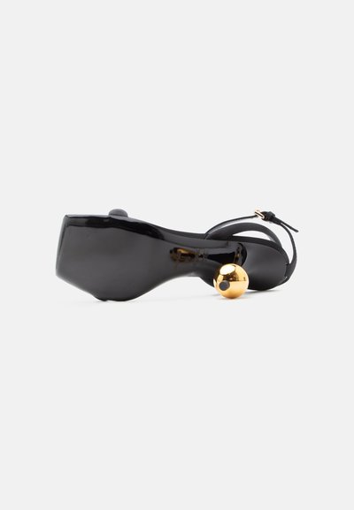 Μαύρο δερμάτινο σανδάλι με τετράγωνη μύτη, λουράκι στον αστράγαλο και χαρακτηριστικό στρογγυλό γυαλιστερό χρυσό τακούνι σε σχήμα μπάλας, φωτογραφημένο από την πλευρά της σόλας.