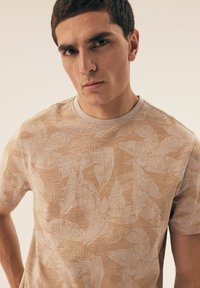 Beżowa koszulka z krótkim rękawem, wykonana z teksturowanej tkaniny, z subtelnym białym wzorem liści i okrągłym dekoltem.