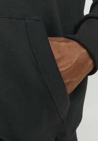 Sudadera negra con puños acanalados. La mano está colocada en un bolsillo lateral, mostrando piel con textura. La tela parece suave y con un diseño casual.