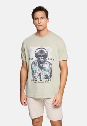 Hombre vestido con camiseta de color beige claro con un gráfico de un gorila con gafas de sol, auriculares y camisa tropical, combinado con pantalones cortos beige claro.