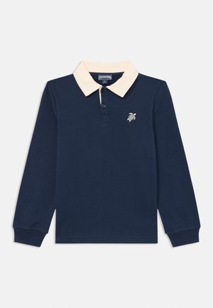 Námořnická modrá polo košile s dlouhým rukávem a světlým béžovým límcem. Na levé straně hrudníku je logo želvy a na légě jsou tři černé knoflíky.