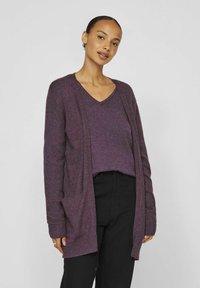 Lila stickad cardigan med öppen front, långa ärmar och två framfickor, lager på lager över en matchande lila v-ringad topp och svarta byxor.