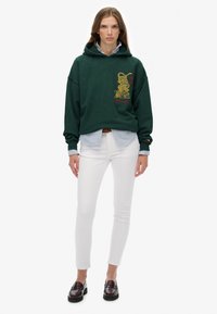 Haut à capuche vert avec un graphisme de dragon et du texte rouge, superposé sur une chemise rayée bleu et blanc ; associé à un pantalon blanc et des mocassins foncés.