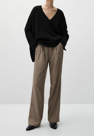 Person wearing loose black V-neck sweater, brown wide-leg trousers, and black pointed shoes, standing with hands in pockets.