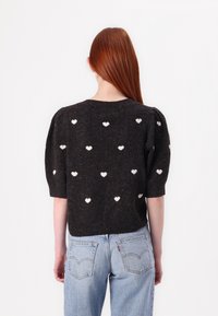 Pull noir en tricot avec manches courtes bouffantes, orné de motifs de cœurs blancs sur toute la surface. Associé à un jean en denim bleu clair.