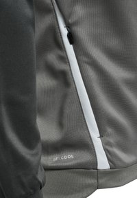 Close-up van een grijze jaszoom en zijzak met een zwarte ritssluiting en witte bies, met het "BEECOOL" logo vlak bij de zoom.