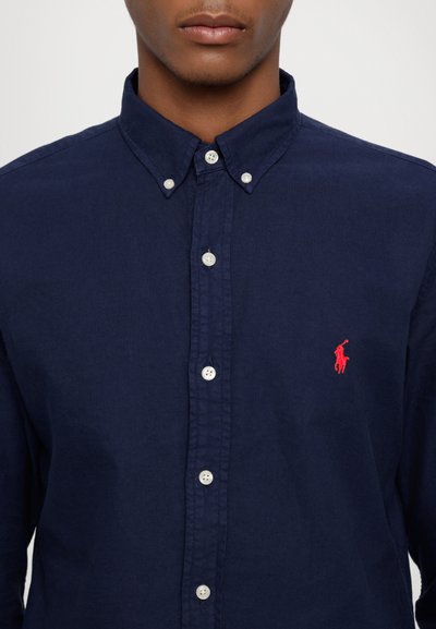 Marineblauwe hette-overhemd van stof met een gestructureerde afwerking, voorzien van witte knopen en een klein rood geborduurd logo op de borst.