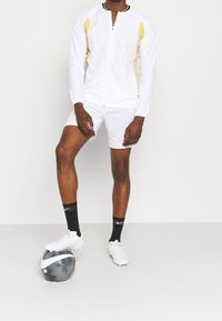 Männlicher Fußballspieler in weißer und gelber Ausrüstung, mit schwarzen Nike-Socken und weißen Fußballschuhen, steht mit dem Fuß auf einem schwarz-weißen Nike-Fußball.