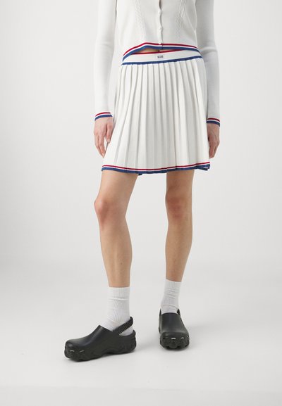 Jupe plissée blanche avec une taille rayée rouge et bleue, associée à des sabots noirs et des chaussettes blanches à la cheville. Cardigan blanc tricoté avec des boutons.