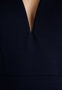 Φόρεμα ναυτικού μπλε με βαθύ V-ντεκολτέ και υφή στο ύφασμα. Το μπούστο έχει ελαφρές πτ�υχώσεις κάτω από το ντεκολτέ και δομημένη σχεδίαση.