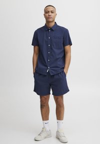 Camisa de botones de manga corta en color navy con tejido texturizado y bolsillo en el pecho, combinada con pantalones cortos a juego. Se lleva con zapatillas blancas y calcetines cortos.