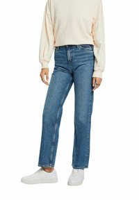 Jeans de pierna recta azul claro hechos de mezclilla con cintura alta, que presentan bolsillos frontales y un ligero desgaste, combinados con zapatillas blancas.