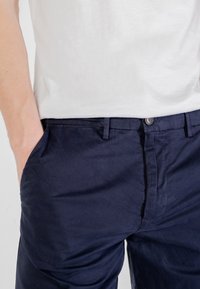 Cino in cotone blu navy con tasche laterali, frontale piatto e chiusura con bottone. Il tessuto ha una consistenza liscia, adatta per un abbigliamento casual.