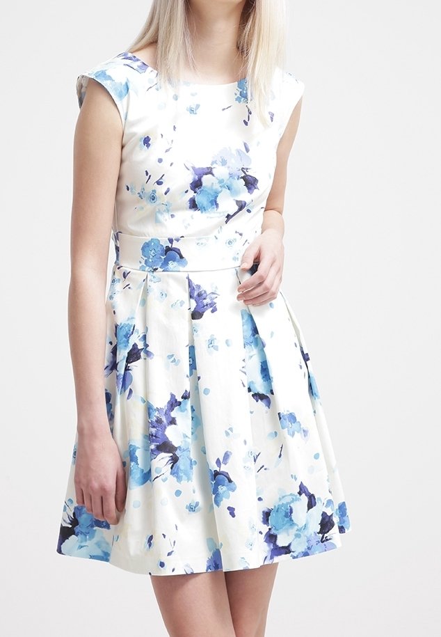 Femme portant une robe blanche sans manches avec un imprimé floral bleu et une jupe plissée, debout devant un fond clair uni.