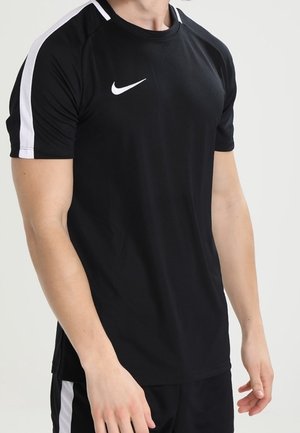 Czarna sportowa koszulka z białymi paskami na ramionach i logo Nike na piersi, noszona przez stojącą osobę z rozluźnionymi ramionami.