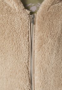 Beige Kunstpelzjacke mit Reißverschluss vorne. Weiche Textur, gerippter Kragen und subtiler Glanz durch die Reißverschlussbeschläge.