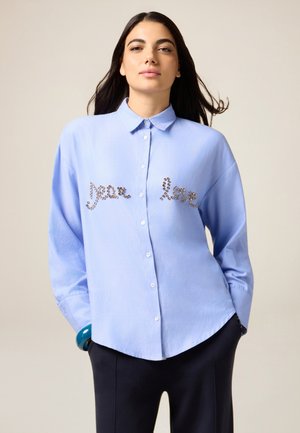 Himmelblaues Hemd mit langen Ärmeln, verziert mit Kristallen, die "Frieden" und "Liebe" über die Brust schreiben.