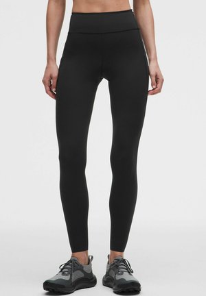 Kobieta ubrana w czarne legginsy z wysokim stanem i szare buty sportowe stoi na jednolitym tle.