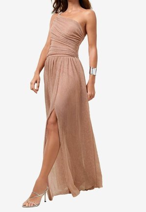 Femme portant une robe de soirée plissée scintillante, asymétrique, couleur or rose, avec une fente haute à la cuisse et des sandales à talons hauts argentées.