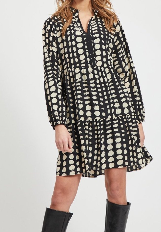 Robe noire à manches longues avec un motif de points blancs irréguliers, jupe à volants, portée avec des bottes noires montantes jusqu'aux genoux.