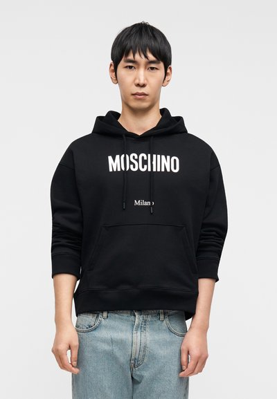 Μαύρο φούτερ με κουκούλα με λευκή εκτύπωση "MOSCHINO" και "Milan". Διαθέτει μπροστινή τσέπη και ριμπ μανσέτες, συνδυάζεται με ανοιχτόχρωμο μπλε τζιν παντελόνι.