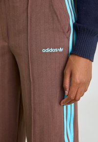 Braune Nadelstreifenhose mit einem türkisfarbenen Seitenstreifen und einem Adidas-Logo in hellem Blau. Weicher Stoff und lockere Passform.