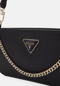 Sac à main en cuir noir texturé avec une bandoulière chaîne dorée et une plaque logo triangulaire portant l'inscription "GUESS USA" au centre.