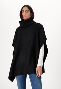 Poncho noir en tricot avec un col haut et côtes. Présente une coupe ample, des manches courtes et une texture qui complète un jean en denim.