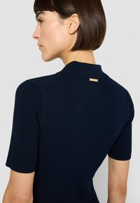 Tumši zilas krāsas ribveduma džempera topiņš ar īsām piedurknēm un apkakli, kakla aizmugurē ar nelielu zelta akcentu.