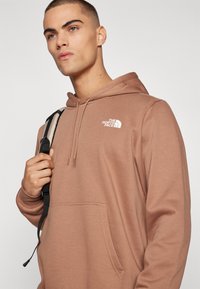 Brauner Kapuzenpullover mit einer frontalen Tasche und weißem Logo auf der Brust. Verfügt über eine Zugkordelkapuze und gerippte Bündchen. Glatte Textur.