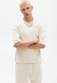 Lichtbeige, kortemouwen mesh top met een V-hals, met een subtiele textuur en een klein logo. Gecombineerd met bijpassende shorts met elastische tailleband.