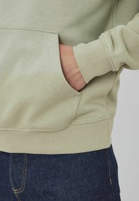 Sweat à capuche vert menthe avec une poche avant et des poignets côtelés, assorti à un jean en denim foncé avec des coutures jaunes et une poche sur la couture latérale.