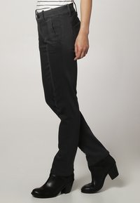 Pantalones stretch negros con corte ajustado, que presentan bolsillos frontales, una textura suave y detalles cosidos. Combinados con botas de tacón negras.