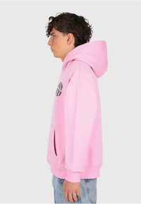Junger Mann im Profil, der einen pinken Hoodie mit schwarzem Text und hellblauen Jeans trägt, vor einem schlichten hellgrauen Hintergrund.