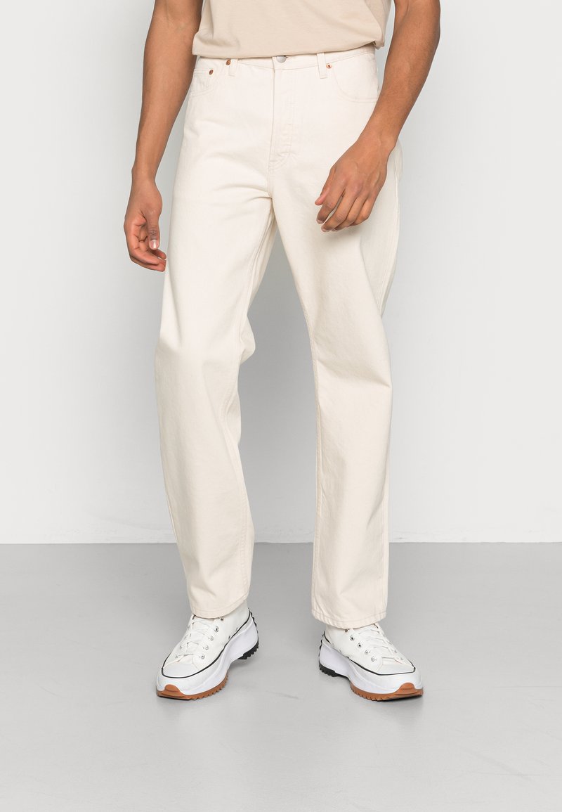 Pantalones de mezclilla de un beige claro con un corte regular, que presentan un diseño de pierna recta y costuras sutiles, combinados con zapatos blancos.