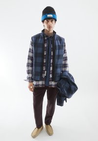 Blauer Karo-Westen über einem gemusterten Hemd, kombiniert mit dunklen Cordhosen und hellen Slippers. Accessoire: schwarze und blaue Mütze.