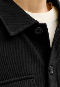 Czarna koszula zapinana na guziki z fakturowanej tkaniny, z kołnierzykiem, czarnymi guzikami i kieszenią na piersi, noszona na kremowej koszuli.