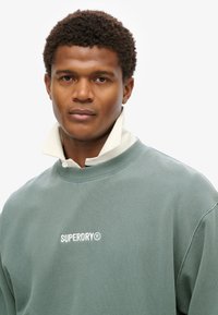 Green sweatshirt with crew neck, ribbed cuffs, and collar layered over it. Features white "SUPERDRY" logo on chest. Smooth texture.
