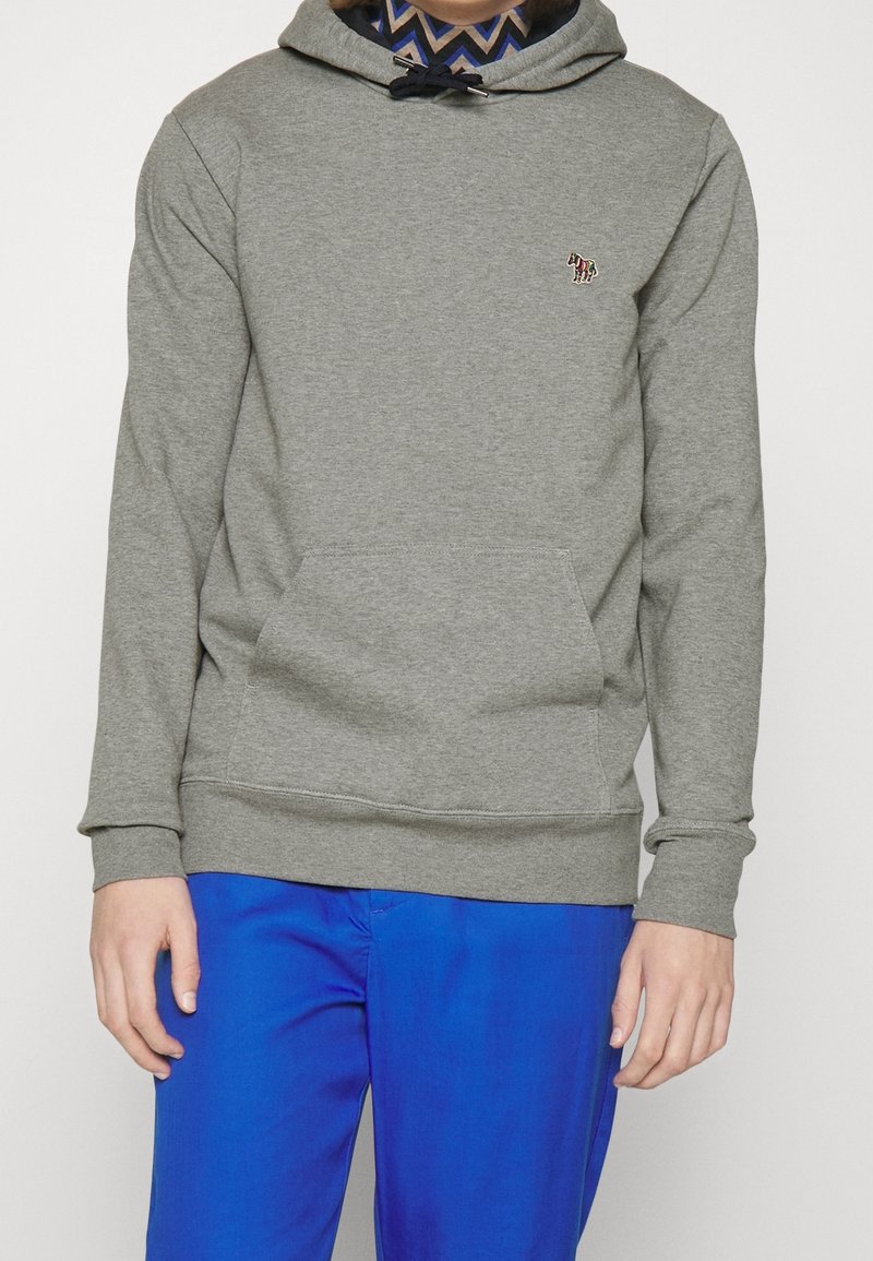 Personne portant un sweat à capuche gris avec un petit logo zèbre brodé sur la poitrine et un pantalon bleu vif.