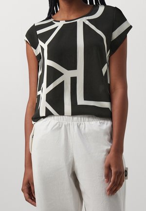 Kobieta ubrana w czarną koszulkę z krótkim rękawem z białymi geometrycznymi liniami, do której dobrano białe spodnie z elastycznym pasem, na jednolitym tle.