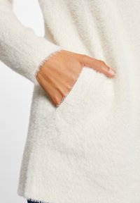 Vit, fluffig tröja med mjuk textur och en framficka. Ärmarna har en rå, lurvig nederkant.