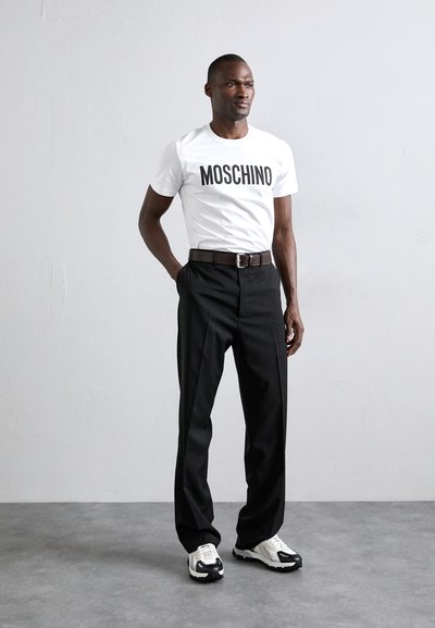 Tricou alb cu logo-ul "MOSCHINO", pantaloni negri, curea maro și adidași negri și albi. Modelul stă pe o suprafață gri, pe un fundal deschis.