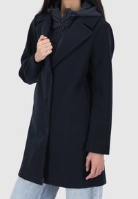 Tmavě modrý kabát s propracovaným střihem, vybavený velkým límcem, knoflíky vpředu a odnímatelnou vycpanou kapucí. Texturovaná tkanina.
