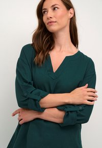 Tyrkysová blúzka s výstrihom do V a trojštvrťovými rukávmi, s ohrnutými manžetami a textúrovanou tkaninou. Má rovný lem a jemné švy.